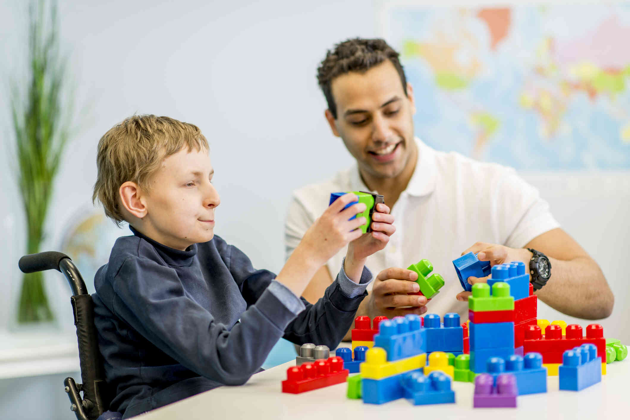 A boy in a wheelchair sits at a table with his dad as they put together jumbo legos.