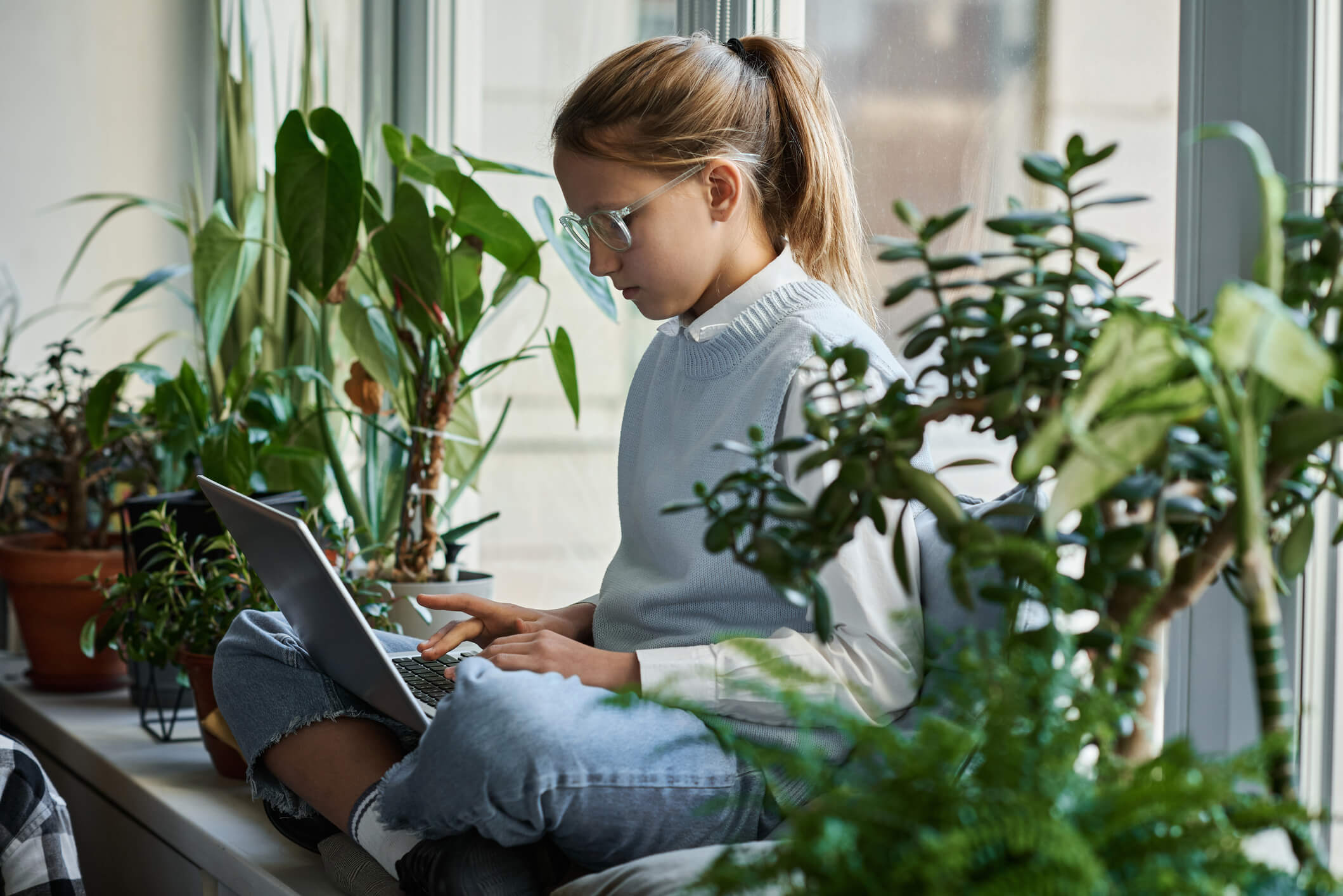 A young girl wearing glasses is sitting on a windowsill and using a laptop; she has a serious expression.